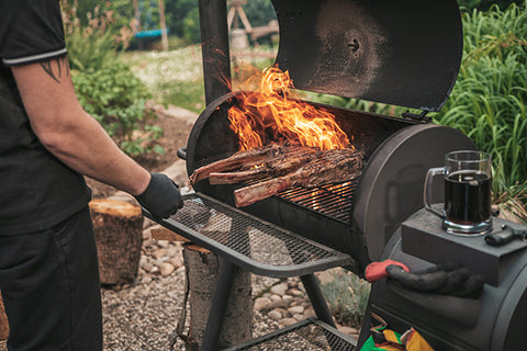 Person grilling with a fire pit and a glass of dark liquid on a stone surface.